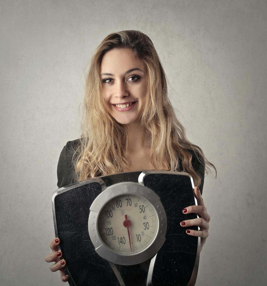 Young woman with blond hair smiling, holding a vintage bathroom scale indoors.