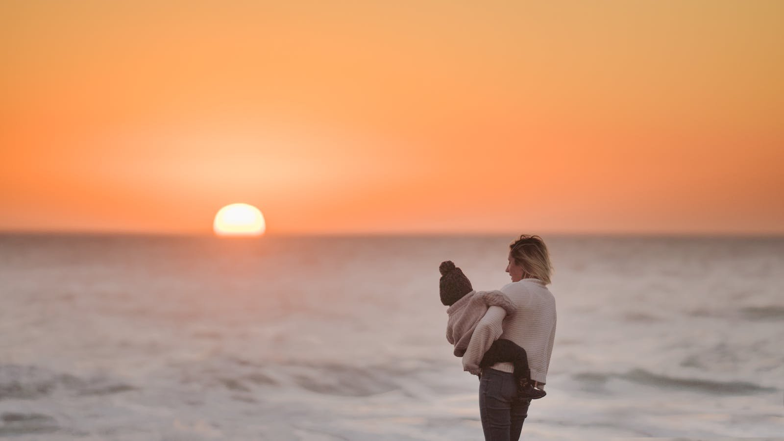 A mother embraces her child while watching the serene sunset by the ocean.