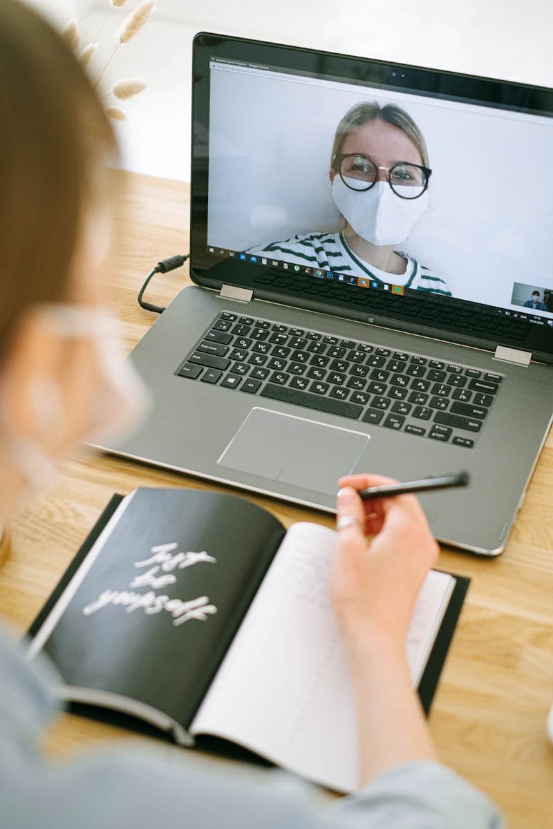 Woman wearing a mask participates in a video call while taking notes in a home office setting.