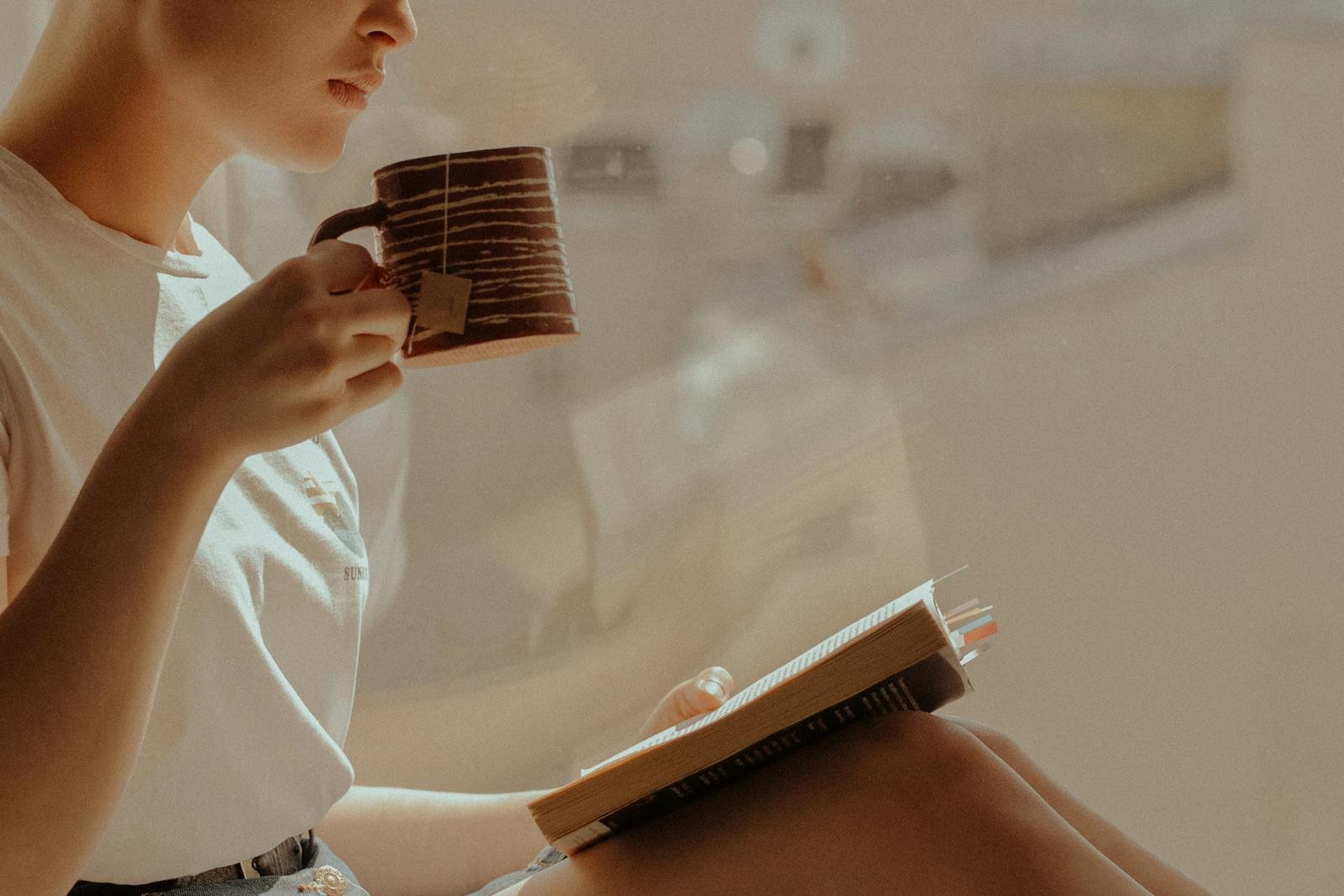 A person enjoys reading a book while sipping tea by a sunlit window, creating a serene atmosphere.