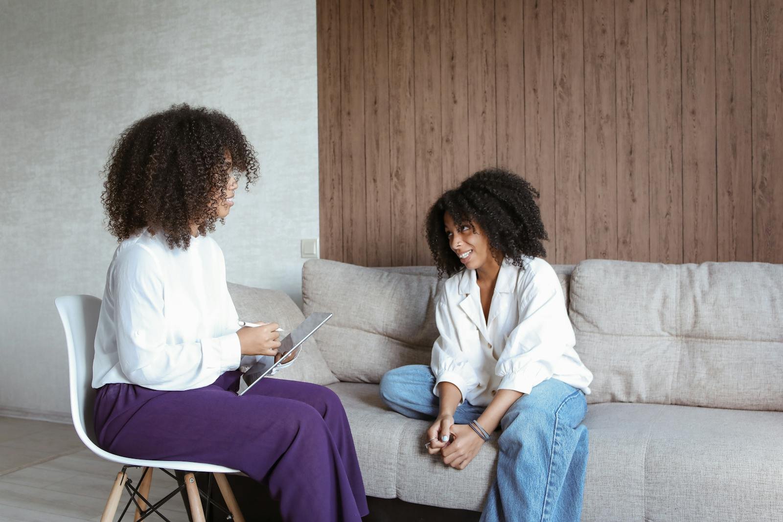 Two women having a conversation in a cozy room, seated on a chair and sofa.