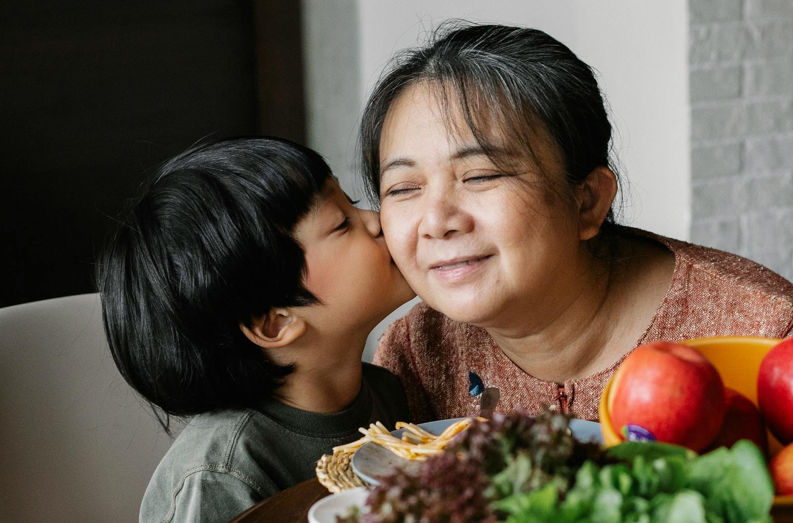 Happy ethnic woman smiling while petting with grandson and sitting at table with various fresh fruits