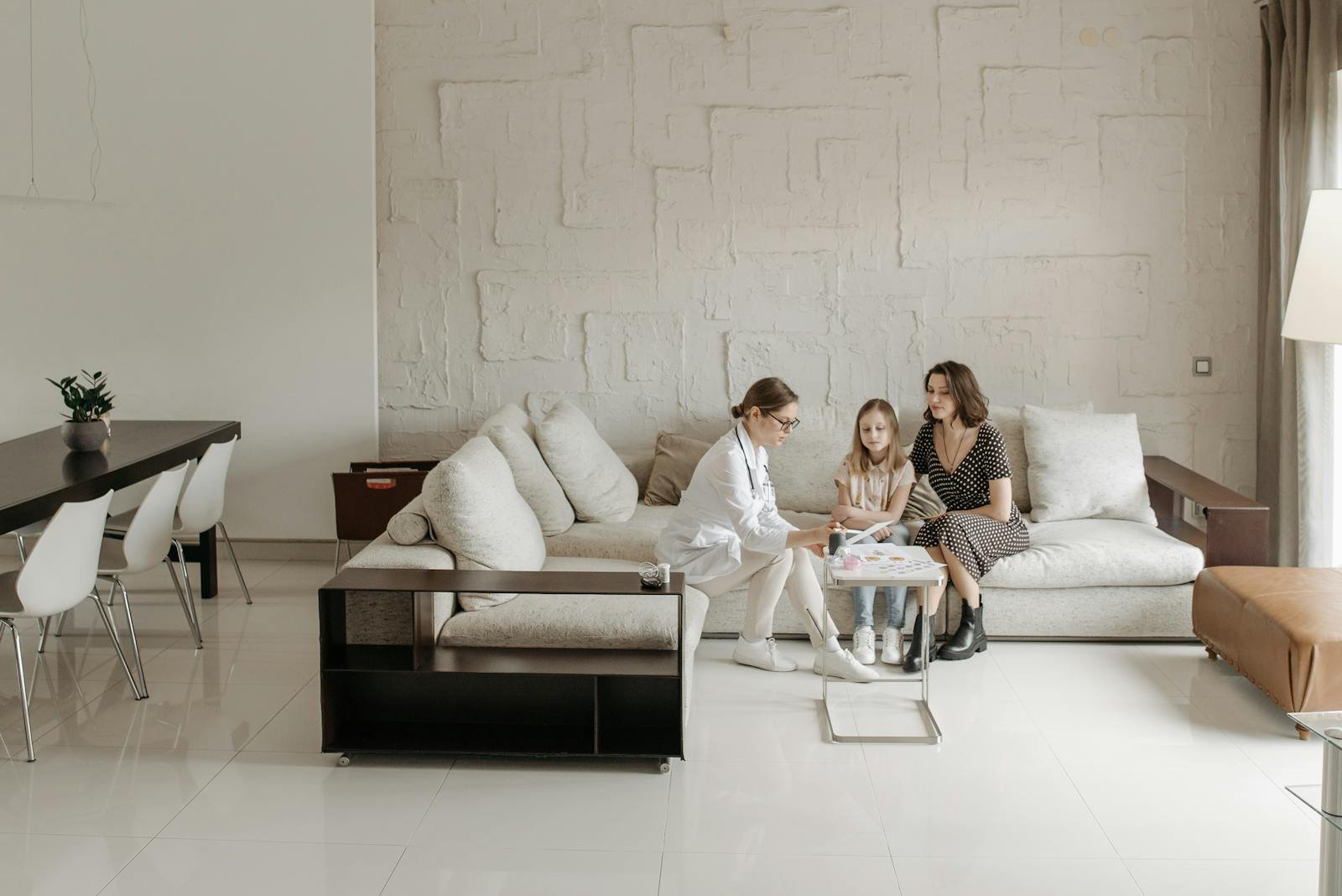 A doctor consults with a mother and daughter in a warm living room setting.