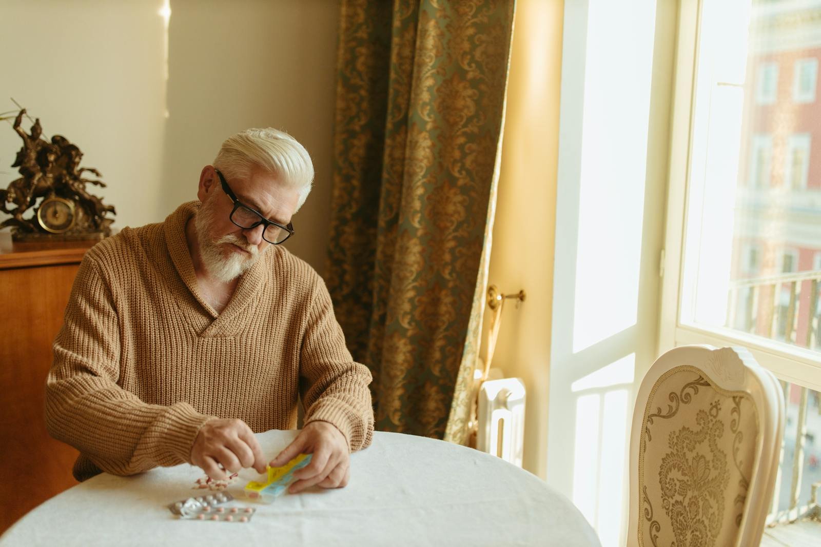 Senior man with gray hair organizing pills at a table by a window, emphasizing daily health routine.