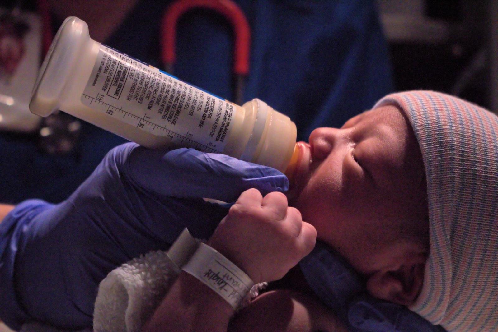 A newborn baby being bottle-fed in a hospital setting by a gloved hand.
