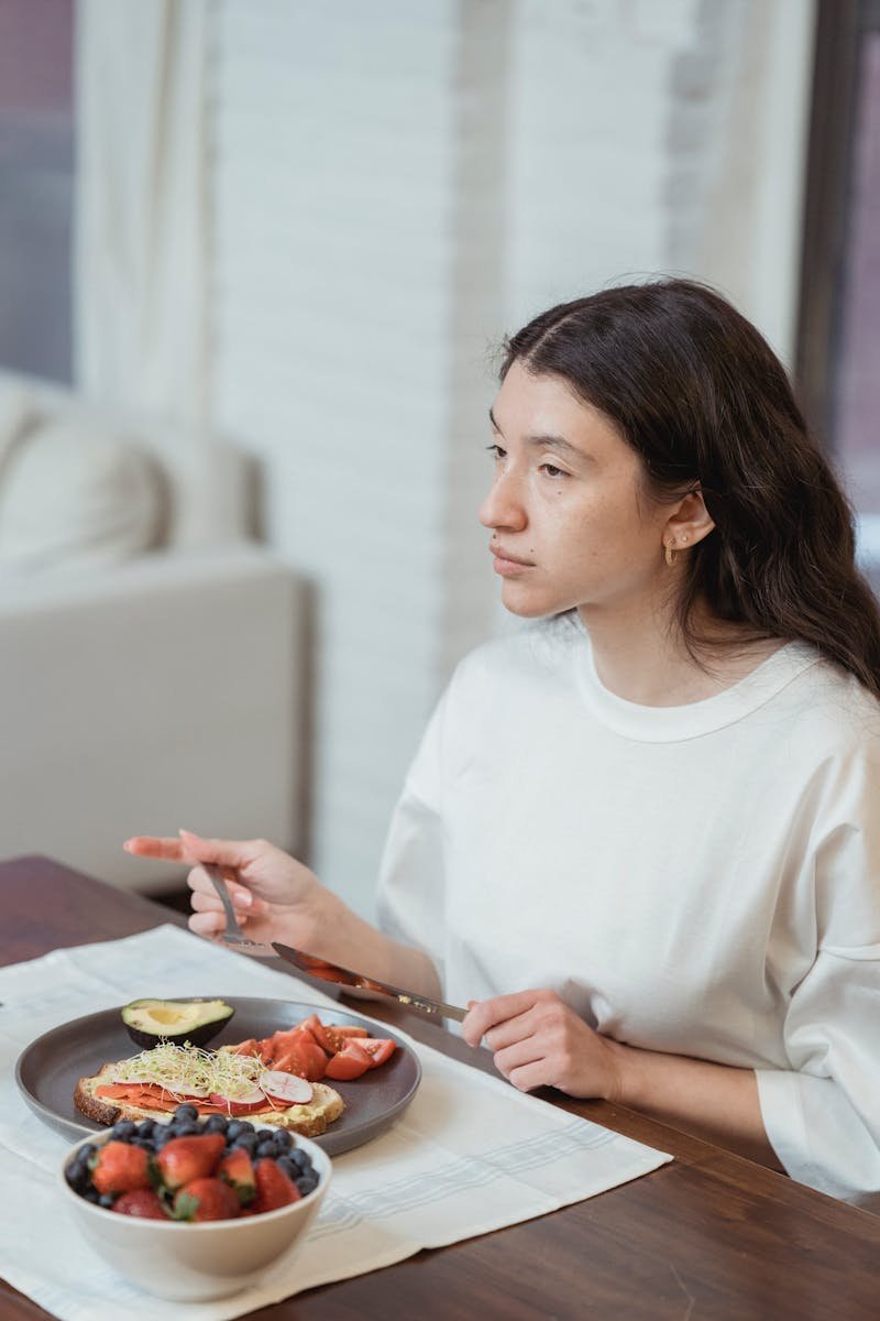 A woman in a white shirt enjoys a healthy breakfast indoors, featuring fruits and toast.
